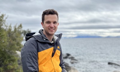 John sits outside on a rocky shore and smiles at the camera