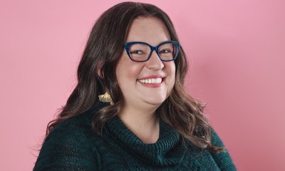 A smiling woman with shoulder-length brown hair, blue glasses, and pale skin.